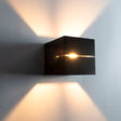 Moderne schwarze Wandleuchte in Würfelform mit offenem Schlitz in der Mitte, die warmes Licht nach oben und unten abgibt – Frontansicht vor einer hellen Wand.