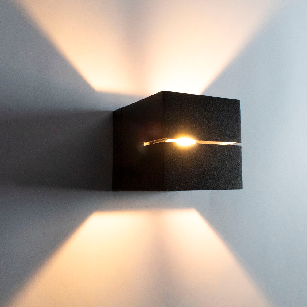 Moderne schwarze Wandleuchte in Würfelform mit offenem Schlitz in der Mitte, die warmes Licht nach oben und unten abgibt – Frontansicht vor einer hellen Wand.