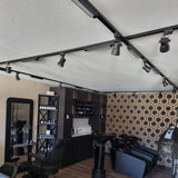 Moderne schwarze 3-Phasen-Schienenbeleuchtung mit verstellbaren Strahlern an der Decke eines Friseursalons, fotografiert von unten mit Blick auf das Innere des Salons und geometrischer Tapete an der Rückwand.