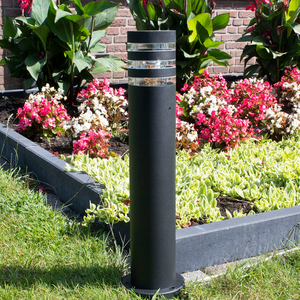 Eine moderne schwarze Gartenleuchte mit rundem Sockel, positioniert in einem farbenfrohen Blumengarten – Vorderansicht bei Tageslicht.