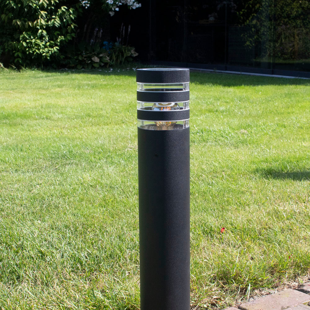 Schwarzer moderner Außenlampensockel in zylindrischer Form mit horizontalem Lichtdiffusor, platziert in einem grünen Garten – Vorderansicht.