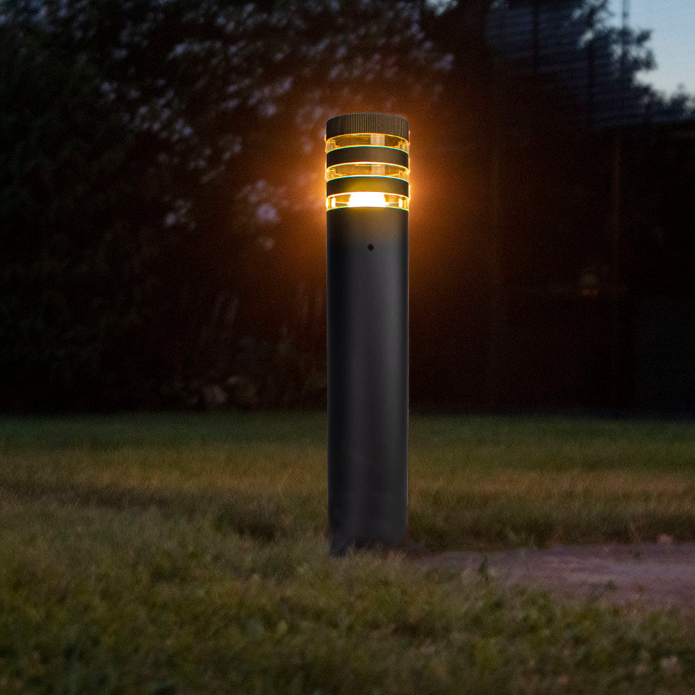 Eine moderne schwarze LED-Außenleuchte mit warmweißem Licht steht in der Dämmerung auf einer Rasenfläche – eine Frontalansicht der stilvollen zylindrischen Gartenleuchte mit horizontalen Lichtöffnungen.