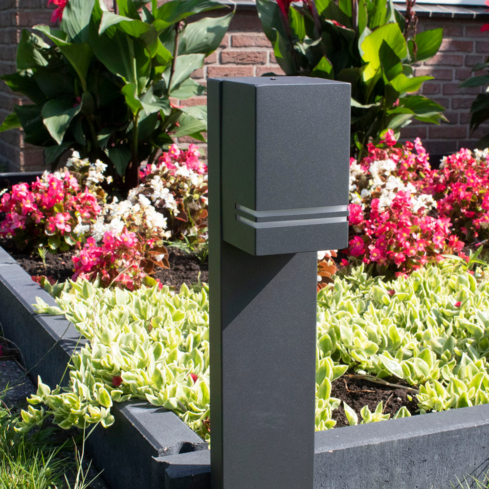 Eine moderne, anthrazitgraue, solarbetriebene Gartenlampe, von der Seite fotografiert, steht entlang eines erhöhten Blumenbeets mit bunten Blumen und Grünpflanzen im Hintergrund.