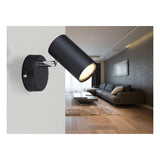 Ein schwarzer, moderner Wandstrahler mit verstellbarem zylindrischen Schirm ist an einer weißen Wand in einem stilvollen Wohnzimmer montiert – Seitenansicht der Lampe mit warmweißem Licht.