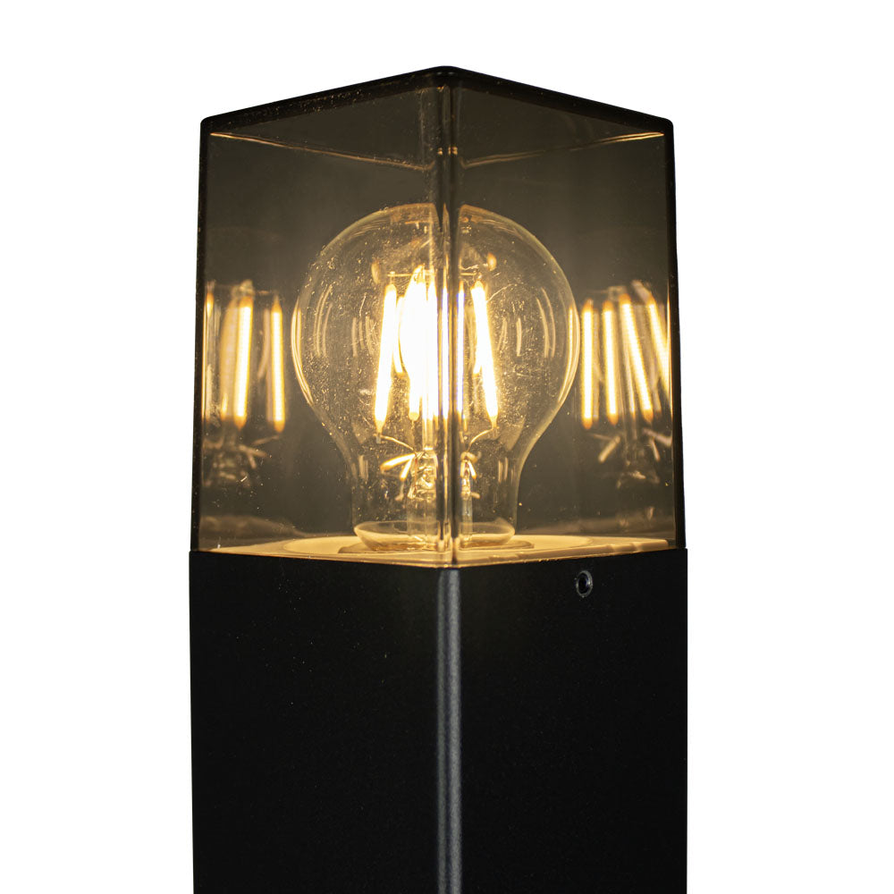 Nahaufnahme einer modernen Außenleuchte mit einer Vintage-LED-Glühbirne in einem transparenten, quadratischen Glasschirm auf einem schwarzen Metallmast – hochwertige Außenstehleuchte für Ihren Garten oder Ihre Terrasse.