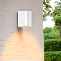 Weiße, moderne Außenwandleuchte mit nach unten gerichtetem Licht, montiert an einer weißen Ziegelwand, Seitenansicht, mit sanfter Beleuchtung an der Wand und einem grünen Garten im Hintergrund.