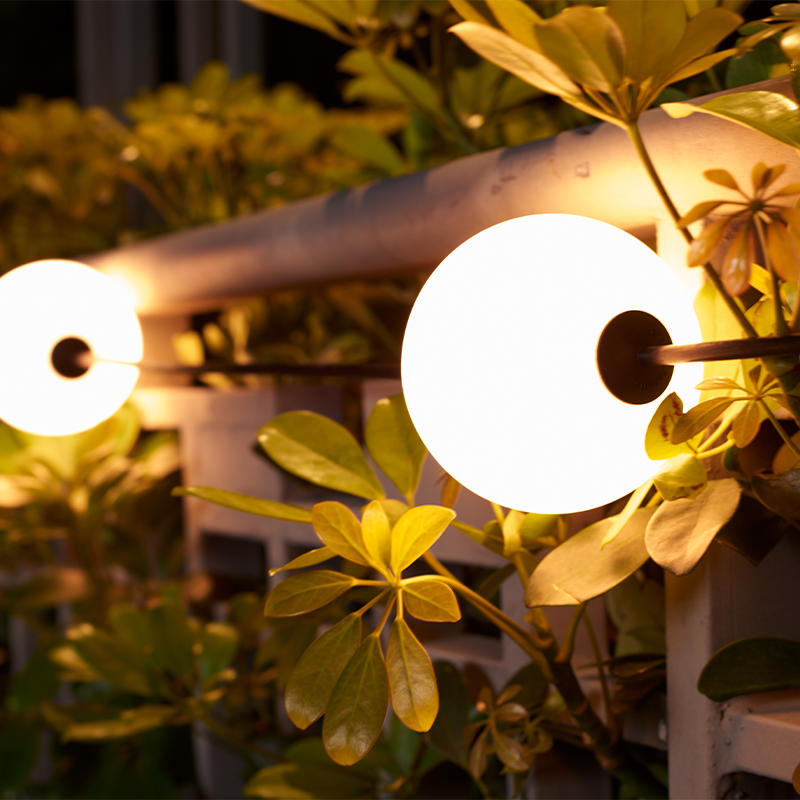 Stimmungsvolle Gartenbeleuchtung: Eine kugelförmige LED-Lampe an einem Balkongeländer befestigt, seitliche Abendansicht mit umgebendem grünen Blattwerk.