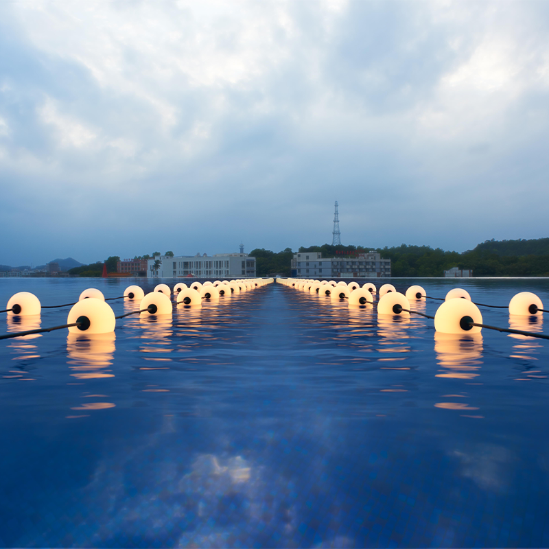 Beleuchtete schwimmende LED-Kugeln in einer Reihe auf der Wasseroberfläche in der Dämmerung, gesehen aus einer niedrigen Frontalansicht in Richtung Horizont mit Gebäuden und einem bewölkten Himmel im Hintergrund.