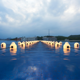 Beleuchtete schwimmende LED-Kugeln in einer Reihe auf der Wasseroberfläche in der Dämmerung, gesehen aus einer niedrigen Frontalansicht in Richtung Horizont mit Gebäuden und einem bewölkten Himmel im Hintergrund.