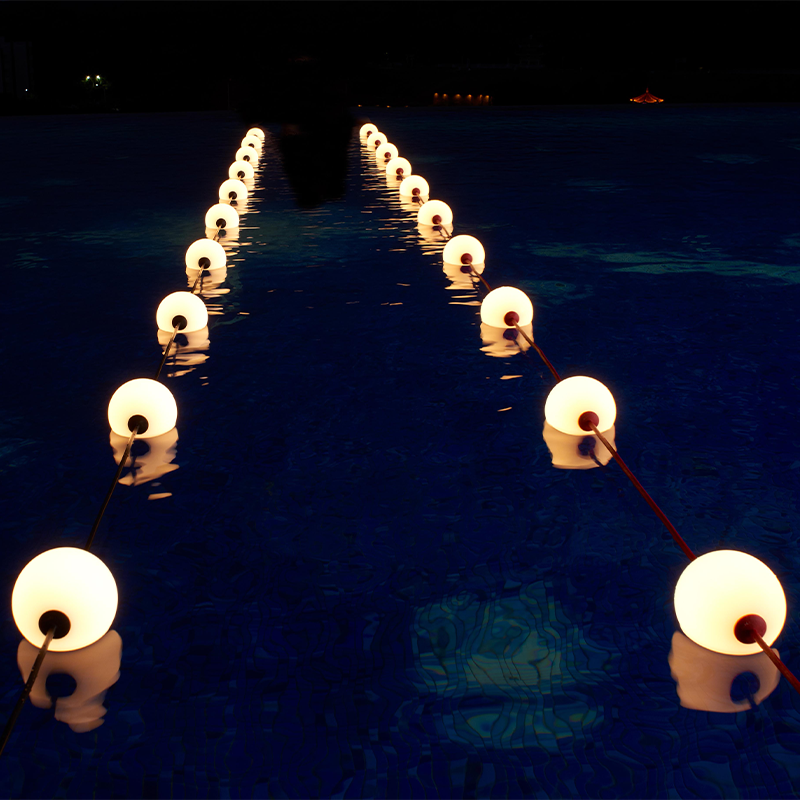 Beleuchtete schwimmende Kugellampen in einer Reihe in einem Schwimmbad bei Nacht, Frontalansicht mit Spiegelung im Wasser.