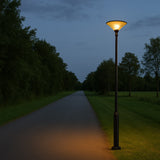 Beleuchtete klassische Straßenlaterne mit konischem Schirm, aufgestellt entlang einer ländlichen Asphaltstraße in der Abenddämmerung, Vorderansicht.