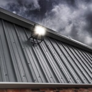 An einer Metallfassade eines Gebäudes montierte LED-Bauleuchte, schräg vor einem bewölkten Himmel betrachtet.