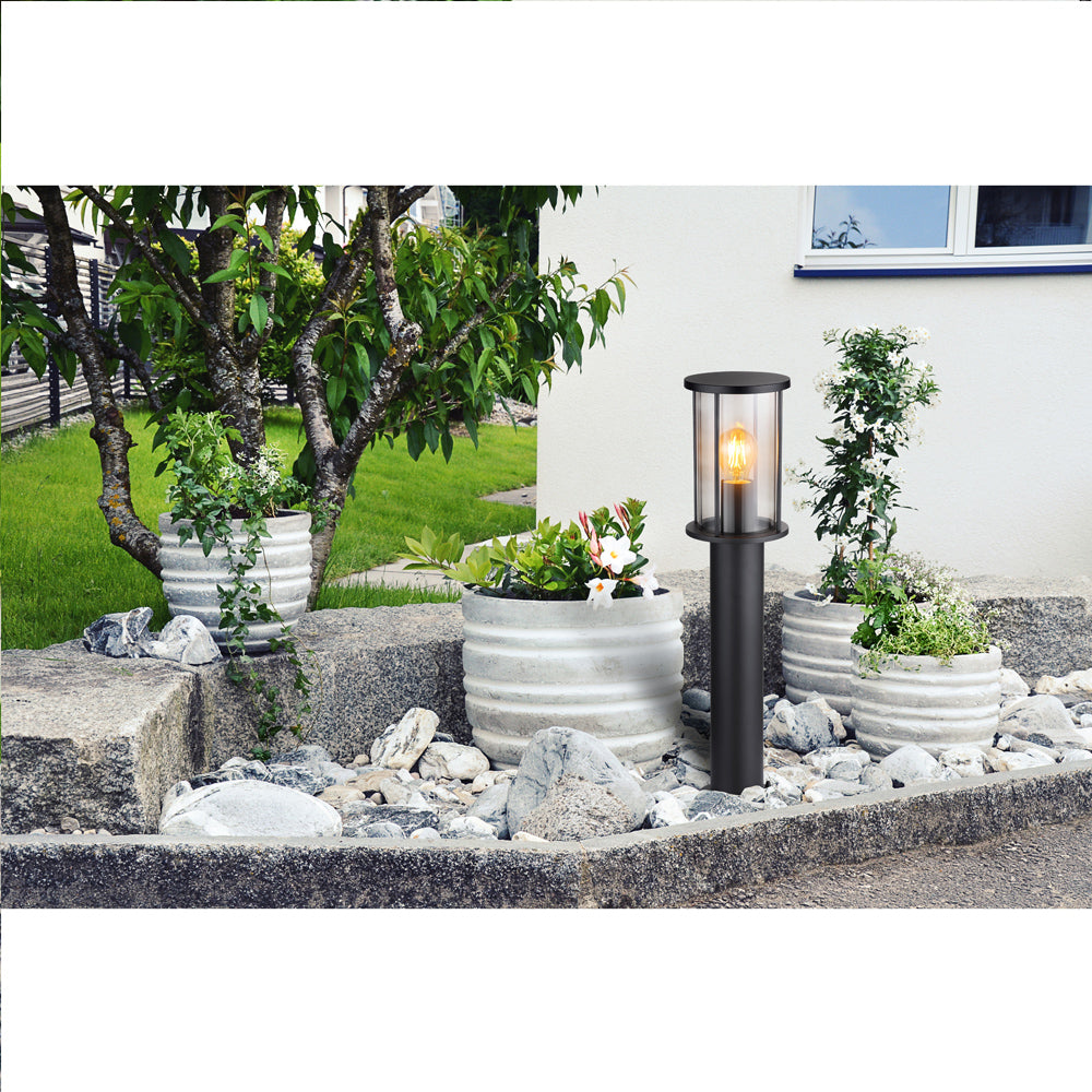 Moderne schwarze Außenlampe, Standmodell, in einer Gartenumgebung, mit einer funktionierenden LED-Glühbirne, umgeben von runden Betonpflanzgefäßen und dekorativen Steinen – Vorderansicht.
