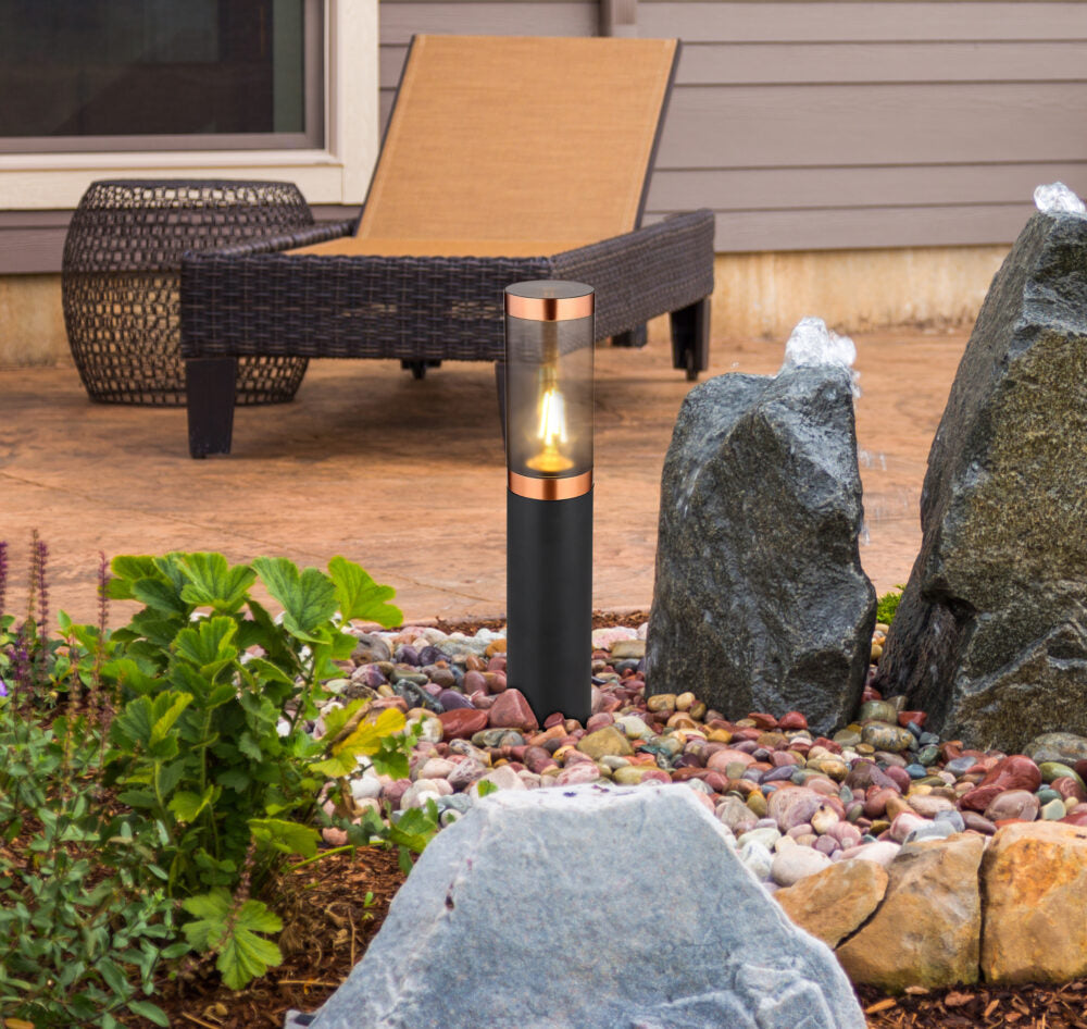 Eine moderne schwarz-kupferfarbene Außenlampe mit zylindrischem Design, in einem Steingarten mit dekorativen Steinen und Grünpflanzen, von der Seite fotografiert, mit einem Liegestuhl auf einer Terrasse im Hintergrund.