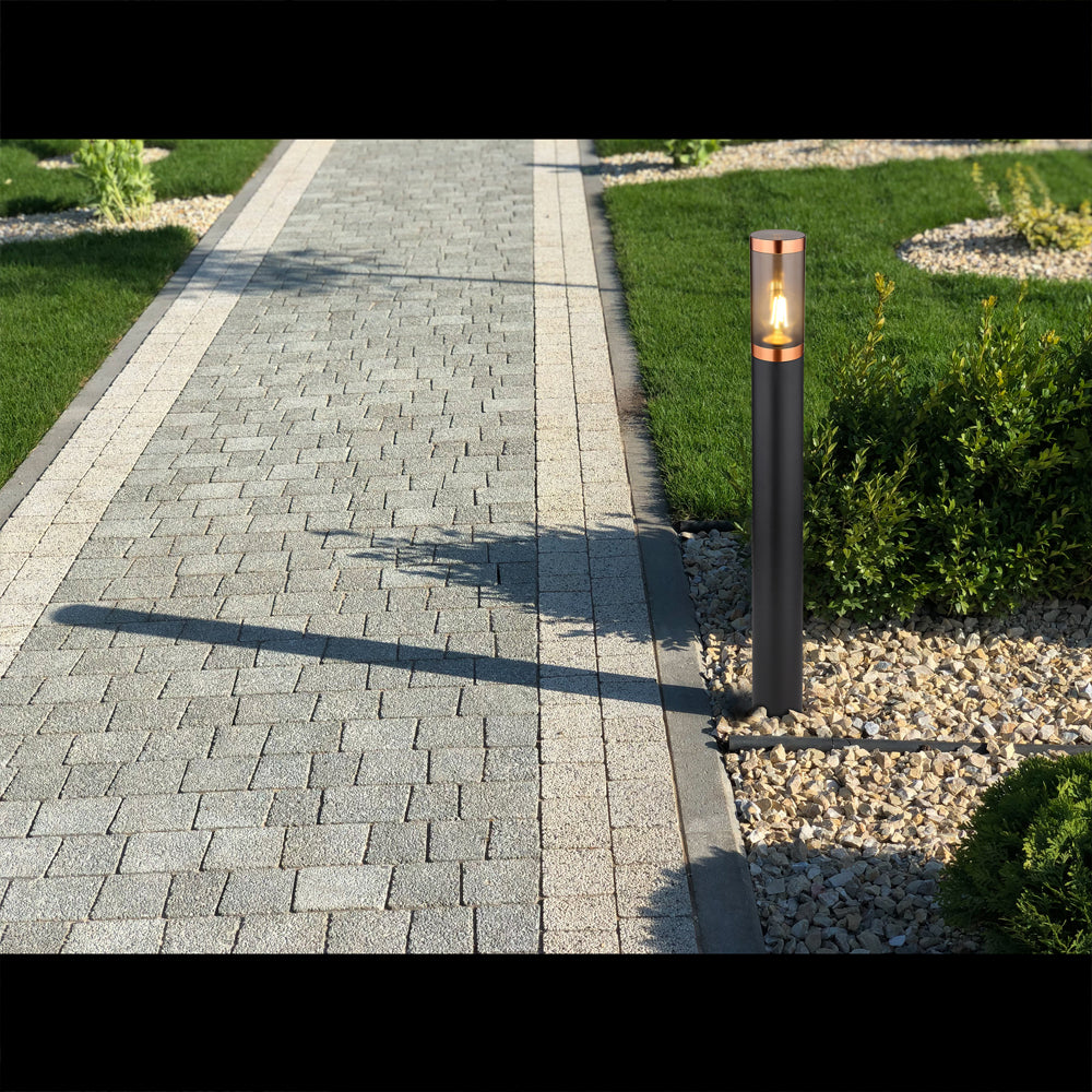Ein moderner schwarzer Außenlampensockel mit kupferfarbenen Akzenten, positioniert entlang eines gepflasterten Gartenwegs mit Gras und Kies – Seitenansicht bei Tageslicht.
