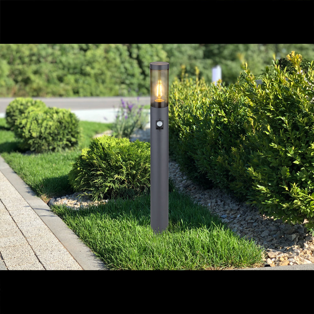 Eine moderne Außenleuchte mit Sensor, die auf einer Rasenfläche entlang eines Gartenweges steht, von vorne fotografiert, im Hintergrund grüne Sträucher.