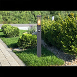 Eine moderne Außenleuchte mit Sensor, die auf einer Rasenfläche entlang eines Gartenweges steht, von vorne fotografiert, im Hintergrund grüne Sträucher.
