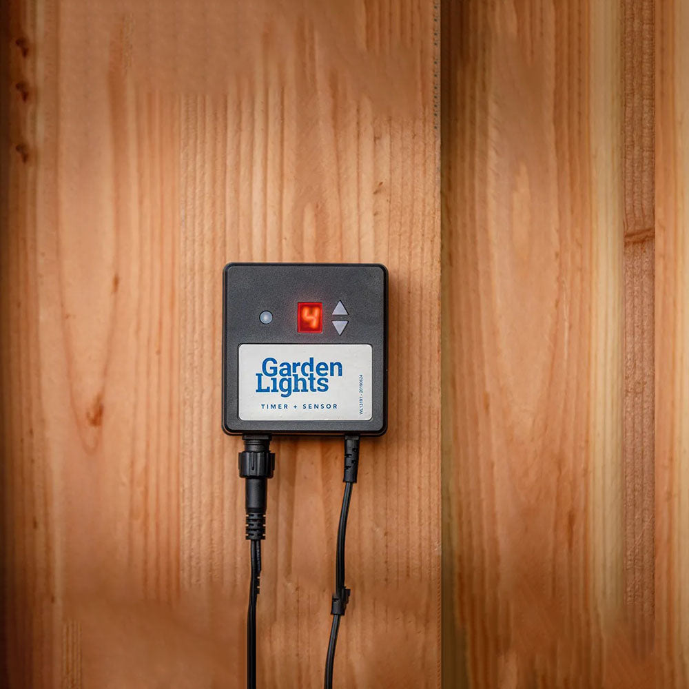 Nahaufnahme eines an einer Holzwand montierten Gartenlicht-Timers mit Sensor, wobei die Digitalanzeige deutlich sichtbar ist und die Stromkabel angeschlossen sind.