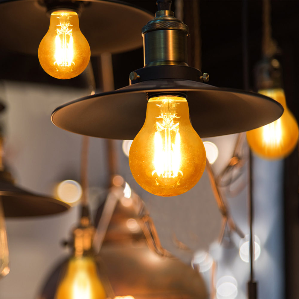 Nahaufnahme einer brennenden Industrie-Pendelleuchte mit einer Retro-Edison-LED-Glühbirne und schwarzem Metallschirm, von vorne vor einem stimmungsvollen, unscharfen Hintergrund fotografiert.
