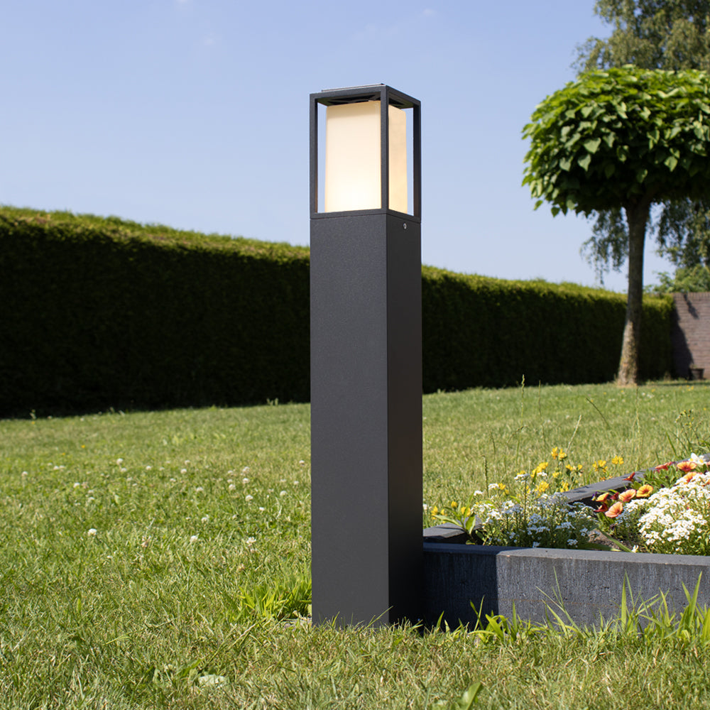 Moderne schwarze Außensockellampe mit quadratischem LED-Laternenschirm, Gartenbeleuchtung auf dem Rasen – schräge Frontansicht bei Tageslicht.