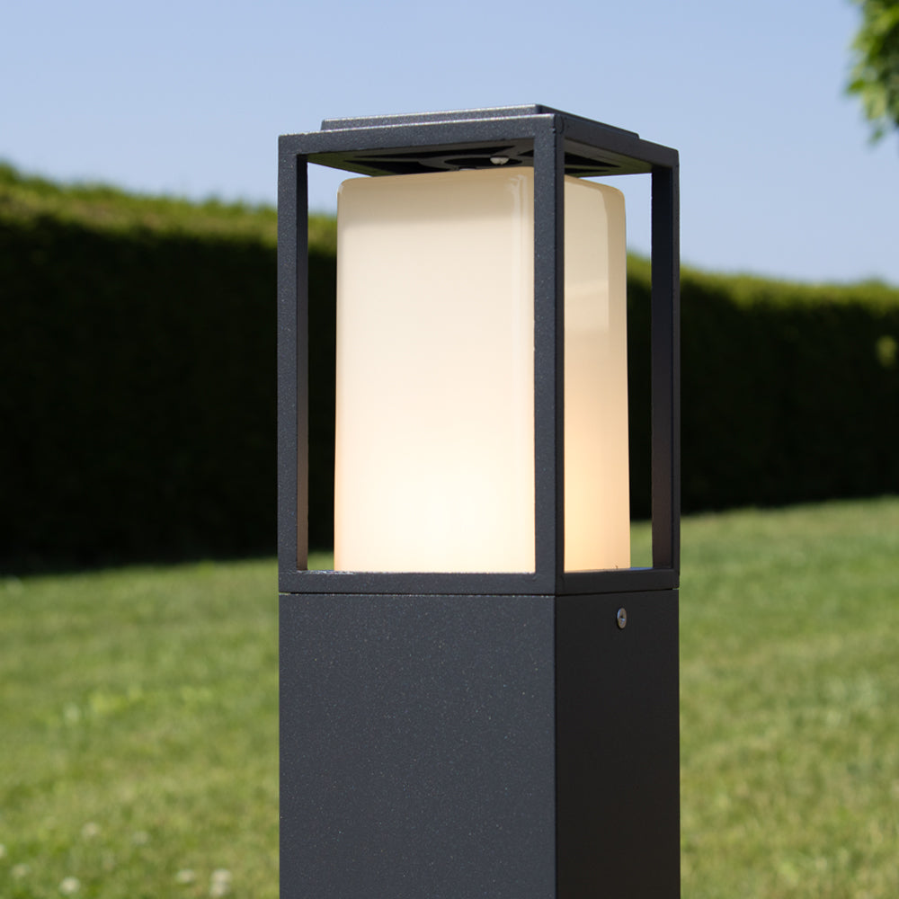 Moderne Außenleuchte mit schwarzem Metallrahmen und mattweißem Leuchtpanel, aus der Nähe und von schräg vorne an einem sonnigen Tag im Garten fotografiert.