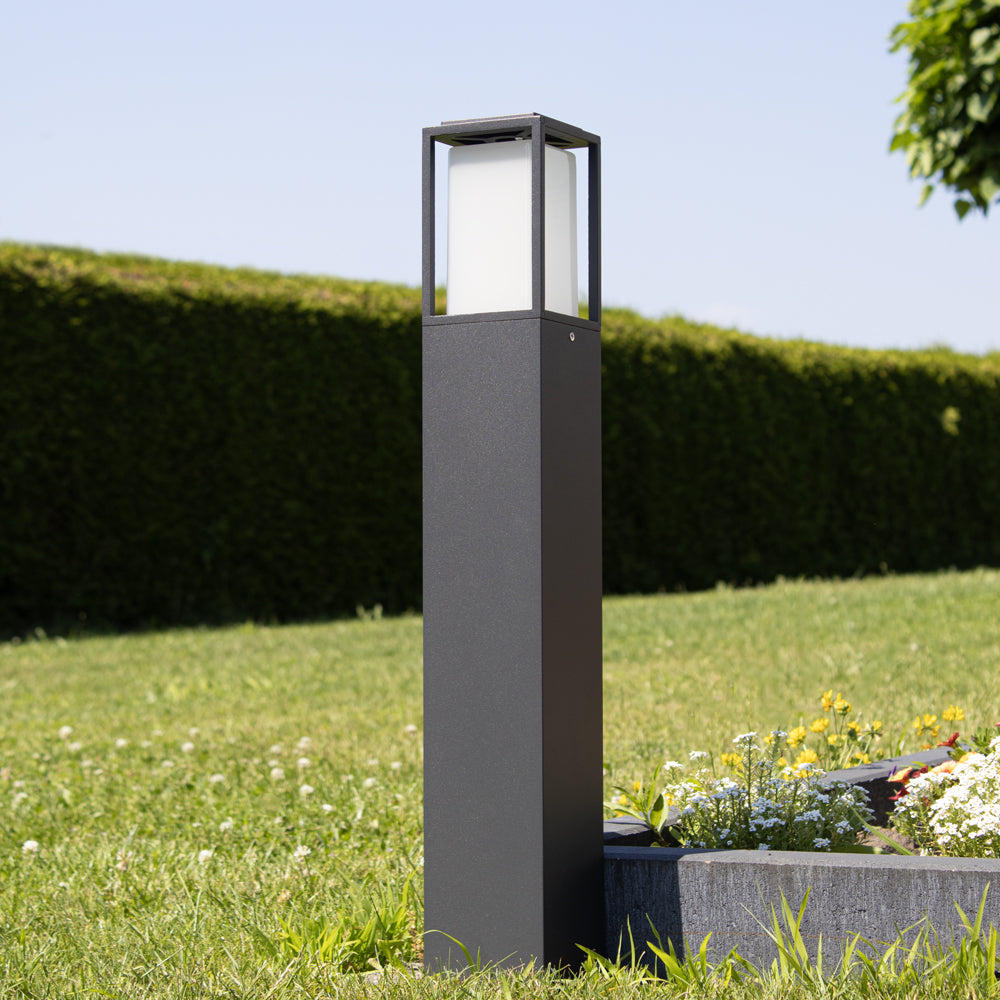 Eine moderne schwarze solarbetriebene Gartenlampe mit quadratischem Laternenschirm, an einem sonnigen Tag von der Seite in einer grünen Gartenumgebung mit einem Blumenbeet im Hintergrund fotografiert.