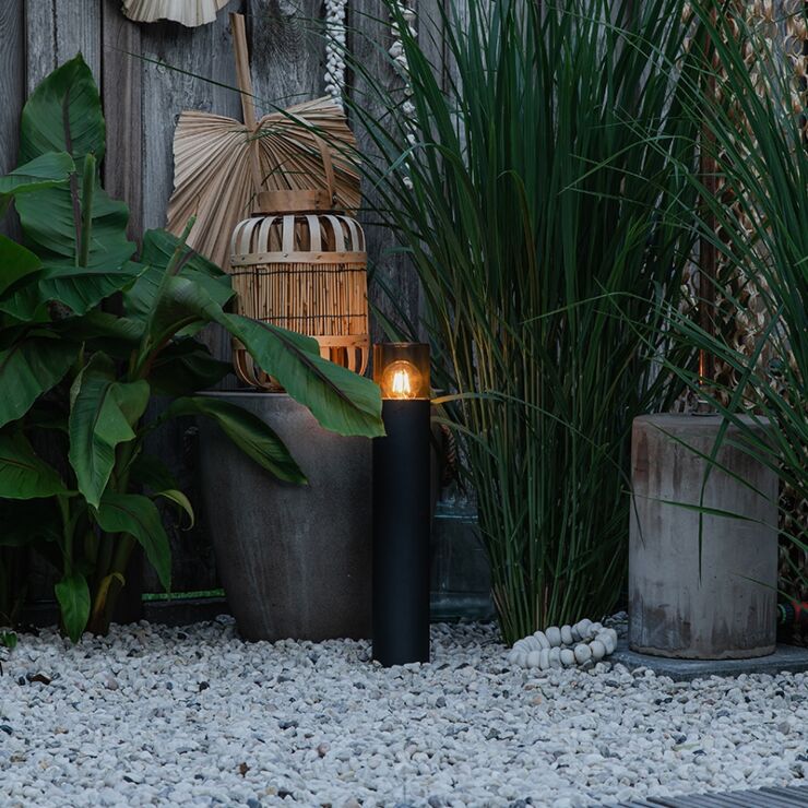 Schwarze, moderne, zylindrische Solar-Gartenlampe, Seitenansicht, platziert in einem stilvoll angelegten Garten mit dekorativem Kies, großen Pflanzgefäßen und Grünpflanzen.