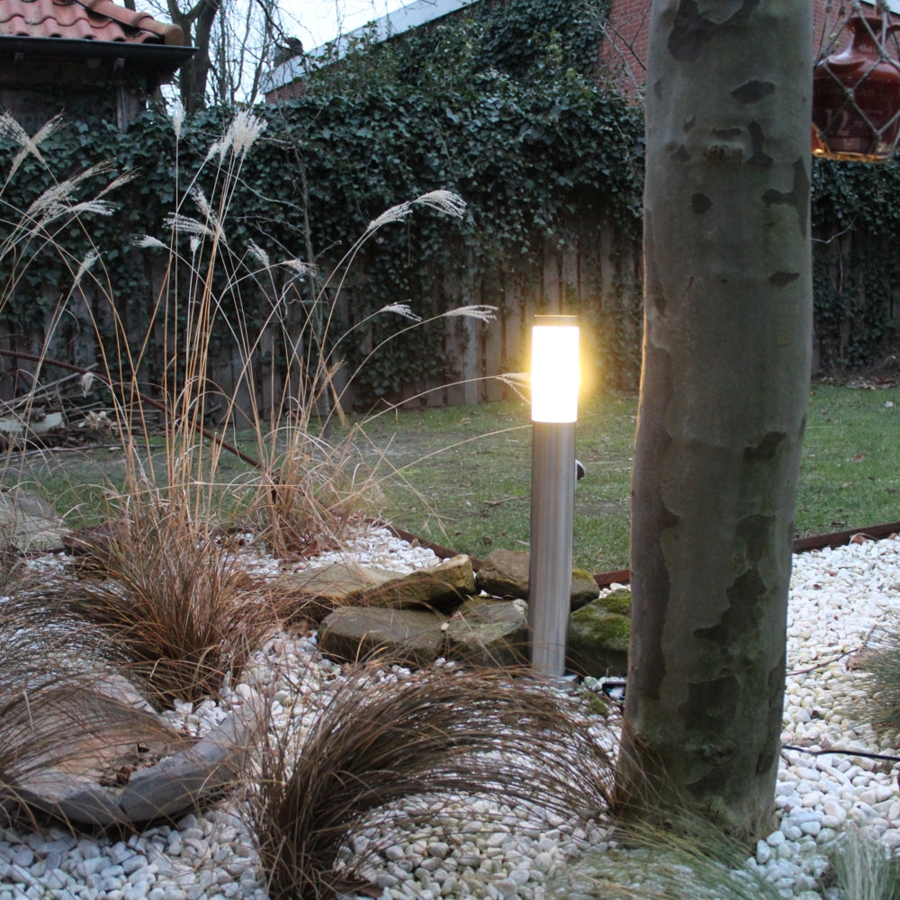 Moderne roestvrijstalen buitenlamp met warm wit licht, geplaatst in een sfeervol aangelegde tuin met siergrind, stenen en siergrassen – frontaal aanzicht bij schemering.