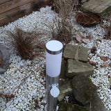RVS buitenlamp met stopcontact, cilindervormig model, staand afgebeeld in een tuin met grind en sierstenen – bovenaanzicht.