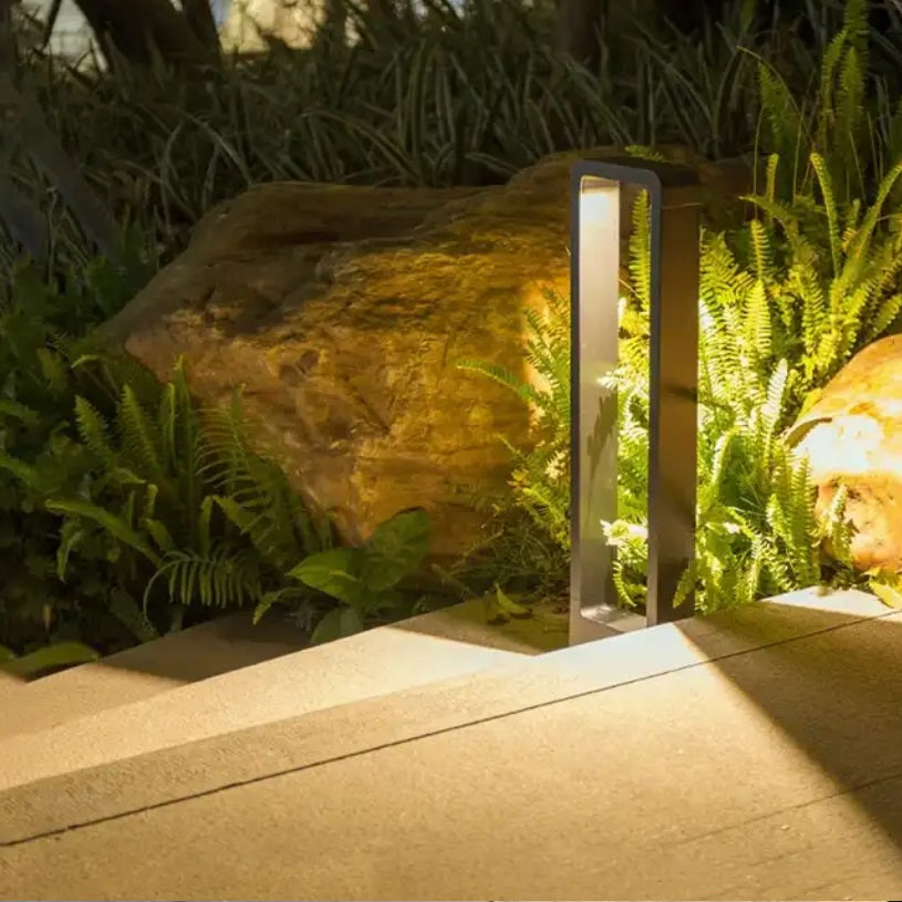 Eine moderne schwarze Gartenpfostenleuchte mit minimalistischem Design beleuchtet einen Weg entlang grüner Farne und eines großen Felsens in einer Gartenumgebung.