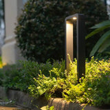 Eine moderne schwarze LED-Gartenleuchte mit rechteckigem, offenem Rahmendesign, die in einem Blumenbeet platziert ist und von oben leuchtet – aus der Perspektive der Dämmerung.