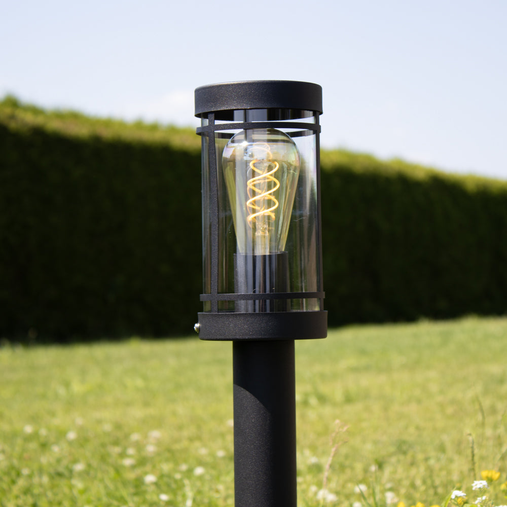 Moderne schwarze Außenleuchte mit sichtbarem Glühfaden in zylindrischer Bauform, von vorne in eine grüne Gartenkulisse gestellt.