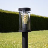 Moderne schwarze Außenleuchte mit sichtbarem Glühfaden in zylindrischer Bauform, von vorne in eine grüne Gartenkulisse gestellt.