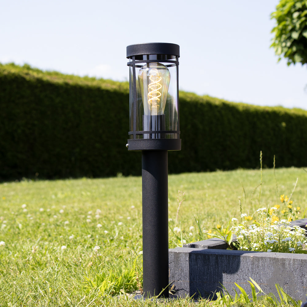 Eine moderne schwarze Außenmastlampe mit klarem Glasgehäuse und sichtbaren Filament-LEDs, entlang einer Rasenfläche in der Nähe eines Blumenkastens positioniert – Frontalansicht.