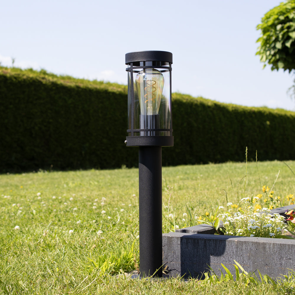 Moderne schwarze Außenmastlampe mit Glasgehäuse und sichtbarer LED-Glühbirne, von vorne betrachtet in einer Gartenumgebung mit Gras und einer blumenreichen Umrandung.