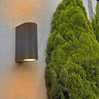 Moderne schwarze Außenwandleuchte mit nach unten gerichtetem Lichtstrahl, montiert an einer hellen Wand neben einem grünen Nadelbaum, Seitenansicht.