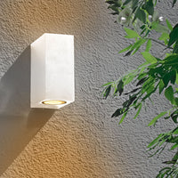 Weiße, moderne Außenwandleuchte, rechteckiges Design mit nach unten gerichtetem Lichtstrahl, Vorderansicht, montiert an einer strukturierten grauen Wand mit grünen Blättern im Hintergrund.