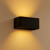 Moderne schwarze Wandleuchte mit Auf- und Ab-Licht, montiert an einer beigen Wand, aus schräger Seitenansicht.