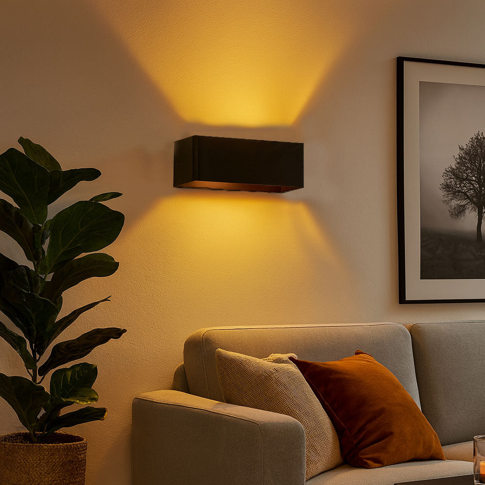 Eine moderne Wandlampe mit warmem, gelbem Licht, montiert an einer beigen Wand in einem gemütlichen Wohnzimmer, mit Blick auf ein graues Sofa, ein dekoratives Kissen, eine grüne Zimmerpflanze und ein schwarz-weißes Kunstwerk – Vorderansicht.