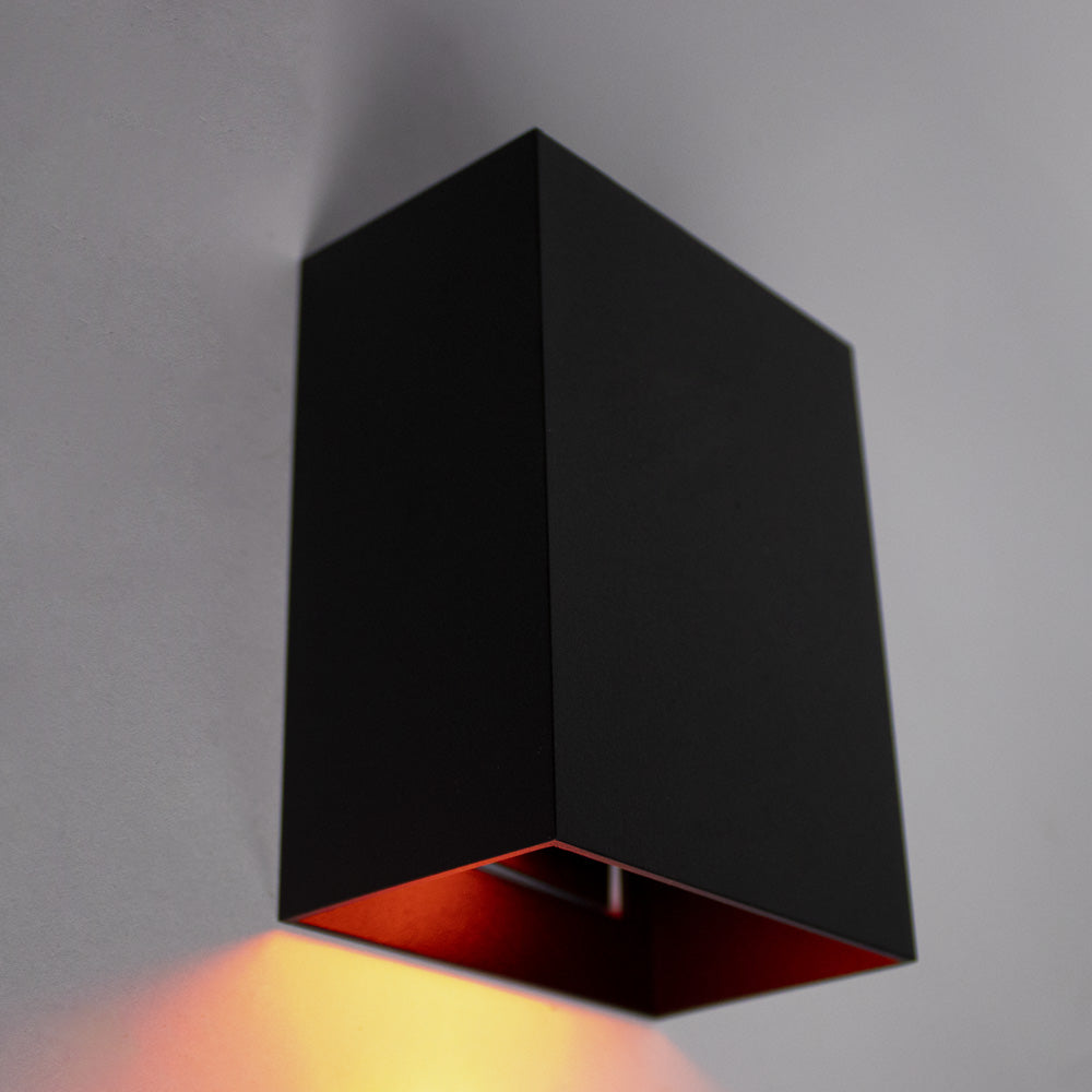 Schwarze, moderne Wandleuchte mit würfelförmigem Design, von schräg unten betrachtet, wobei der warme Lichtstrahl nach unten auf eine graue Wand scheint.