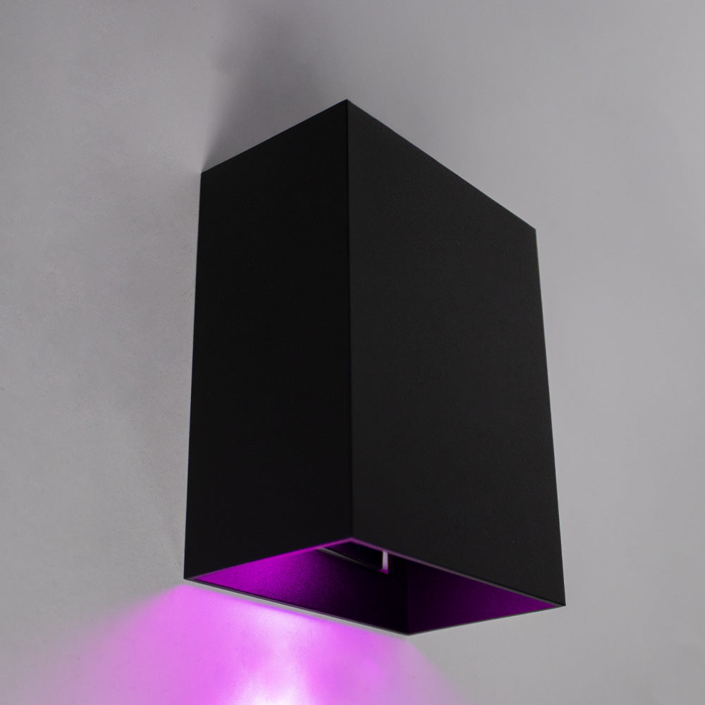 Schwarze moderne Wandleuchte mit Downlight-Effekt und violettem LED-Licht, schräg vor einer grauen Wand betrachtet.