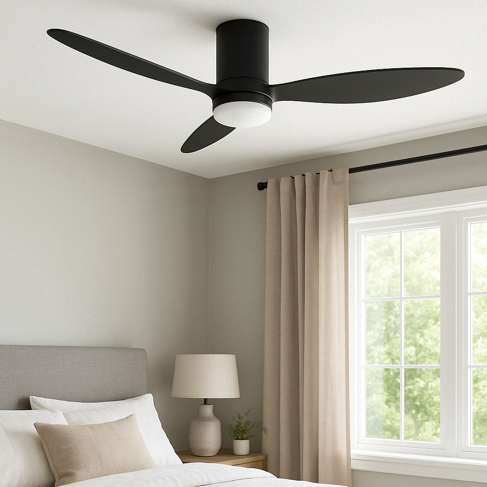 Ein moderner schwarzer Deckenventilator mit drei Flügeln und integrierter LED-Beleuchtung ist an der Decke eines minimalistischen Schlafzimmers montiert – eine Seitenansicht aus einem Winkel, die das Fenster, die Vorhänge und das Bett zeigt.