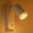 Moderne weiße Wandleuchte mit USB-Anschluss und Schalter, Seitenansicht mit warmer Beleuchtung vor einer beigen Wand.
