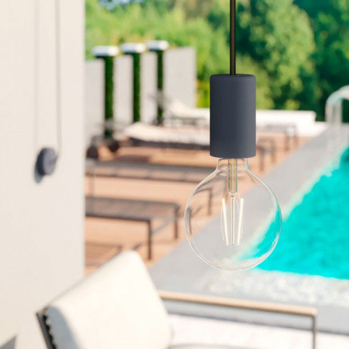 Moderne Pendelleuchte mit schwarzer Fassung und runder LED-Glühbirne, Nahaufnahme, montiert über einem luxuriösen Außenpool mit Sonnenliegen im Hintergrund.