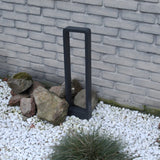 Eine moderne schwarze Außensockellampe mit Metallrahmen, platziert in einem Garten mit weißem Kies und Ziersteinen, von schräg vorne vor einer hellgrauen Ziegelmauer gesehen.