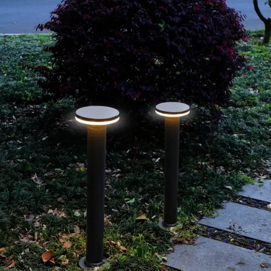 Zwei moderne schwarze Gartenpfosten mit runder LED-Beleuchtung, entlang eines Gartenwegs in abendlicher Umgebung platziert – Frontalansicht.