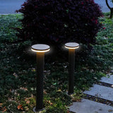Zwei moderne schwarze Gartenpfosten mit runder LED-Beleuchtung, entlang eines Gartenwegs in abendlicher Umgebung platziert – Frontalansicht.