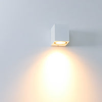 Weiße moderne Wandleuchte, würfelförmiges Modell, mit nach unten gerichtetem warmen LED-Licht, seitlich vor einer weißen Wand fotografiert.