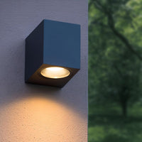 Moderne schwarze Außenwandleuchte in Würfelform mit nach unten gerichtetem warmen LED-Licht, an einer Wand montiert – Vorderansicht.