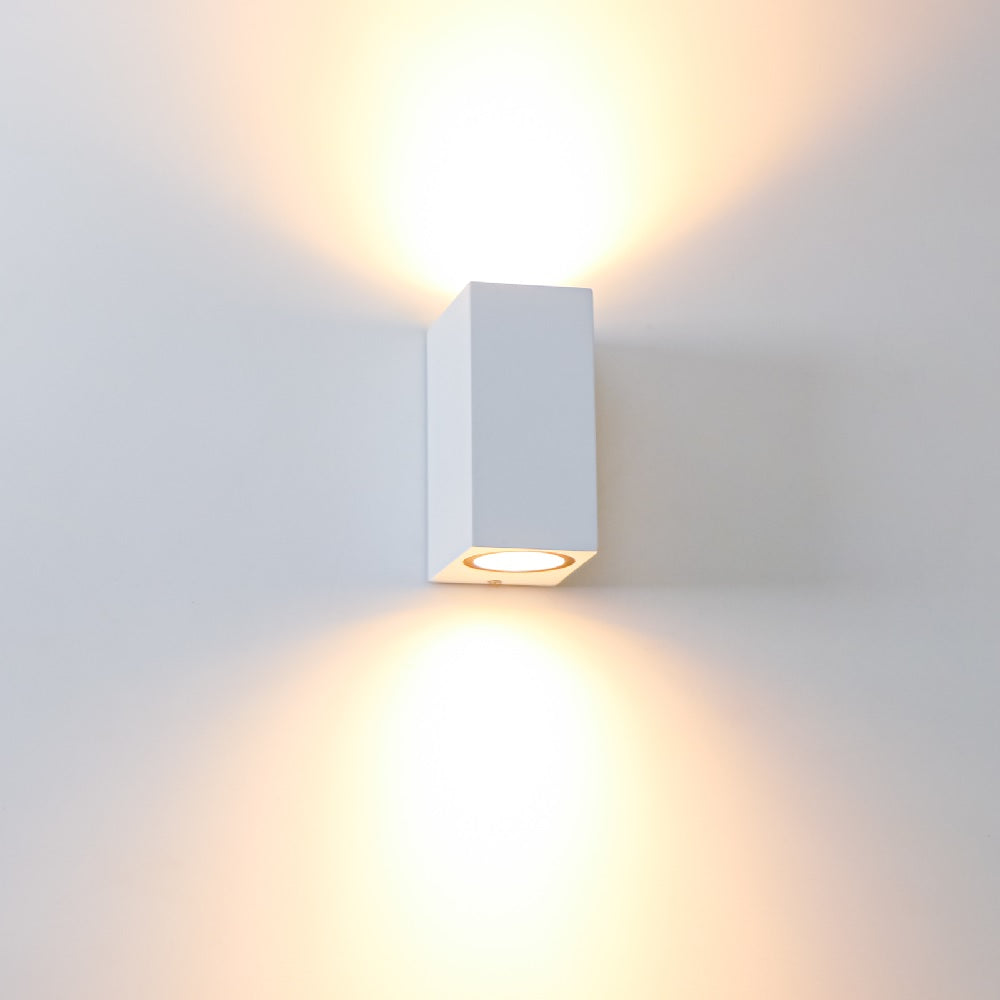 Weiße, moderne Wandleuchte in Würfelform mit Up- und Downlight-Effekt, montiert an einer weißen Wand, Vorderansicht.
